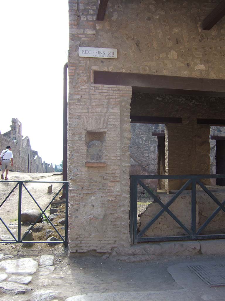 Street shrine or niche outside I.12.5 Pompeii. September 2005.
This is on Via dell’ Abbondanza, at the corner with Vicolo dei Fuggiaschi. 
According to Della Corte, on the red pilaster which ended the insula on the east side, two electoral programmes were found.
The first – 
GAVIVM II VIR.      [CIL IV 7442]
The second was immediately below the first - 
AMPLIATUM L F AED
                          VICINI                                                                                                           
                          SVRGITE ET
                           ROGATE
                           LVTATI F[ac]     [CIL IV 7443]
The text was placed towards the right, because the pilaster was interrupted in the middle by a rectangular niche.
In the niche was fixed a coarse rough stone resembling the outline of a human head, not the usual marble bust, as often seen here and there in the street.
See Notizie degli Scavi di Antichità, 1914, (p.204)
See Epigraphik-Datenbank Clauss/Slaby (www.manfredclauss.de).

According to PPM, inserted in the rectangular niche on the left side of the doorway was a blue lava stone, which had been a “lava-bomb” erupted during a prehistoric eruption and which was supposed to have been attributed with sacred and magical values. 
This was inserted into an apsed rectangular niche, with the lower level consisting of a tile projecting by about 6 cm and closed into the upper area by another tile with architrave under a flat arch in brick. 
See Carratelli, G. P., 1990-2003. Pompei: Pitture e Mosaici. Vol. II. Roma: Istituto della enciclopedia italiana. (p.736)

