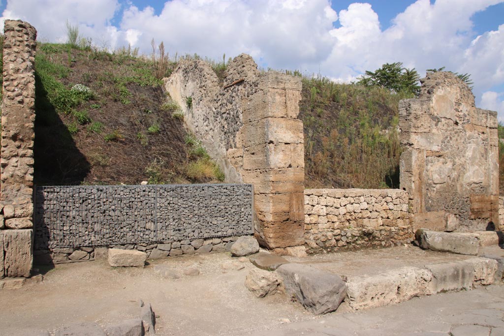 III.7.1, on right, Pompeii. October 2022. 
Entrance doorway on north side of Via dell’Abbondanza, with unexcavated roadway between insulae with grey gabions, on left. 
Photo courtesy of Klaus Heese
