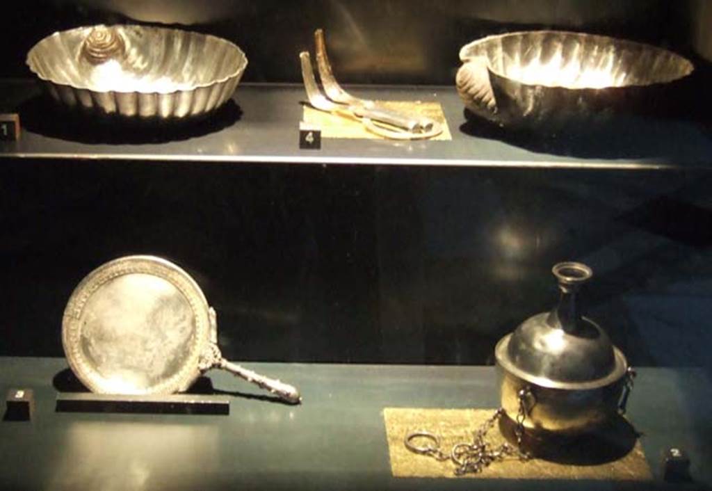 V.1.18 Pompeii. Silver found in the ala “e”. 
Now in Naples Archaeological Museum.
Top left to bottom right:-
Two shell pattern containers. Inventory numbers 110863 and 110864
Between them are two strigili joined by a ring. Inventory number 110862.
Concave circular mirror with handle. Inventory number 110861.
Container with cylindrical neck. Inventory number 110841.
