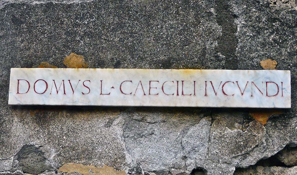 V.1.26 Pompeii. May 2005. Entrance with name plaque DOMUS L CAECILI IUCUNDI.
Many electoral programmes were written on the façade:

[M.]  Holconium  Priscum  II  vir(um)  i(ure)  d(icundo)
d(ignum)  r(e)  p(ublica)  o(ro)  v(os)  f(aciatis)
IVCVNDVS  ROGAT   [CIL IV 3428]
According to Della Corte, this was written to the right of the main entrance doorway at 26.

He also quotes:
[L.]  Ceium  Secundum –  II  vir(um)
Q(VINTVS)  S(EXTVS)  CAECILI   IVCVNDI   ROGAMVS  [CIL IV 3433].
Della Corte says the second programme was found on the pilaster to the right of the next shop at number 27.
This shop was part of the same house.
See Della Corte, M., 1965. Case ed Abitanti di Pompei. Napoli: Fausto Fiorentino. (p.18)

According to Cooley, the following was found on the façade of the house:
We, Quintus and Sextus Caecilius Iucundus, ask for Ceius Secundus as duumvir. [CIL IV 3433 = ILS 6404a]
See Cooley, A. and M.G.L., 2004. Pompeii: A Sourcebook. London: Routledge. (F17 on p. 116)

