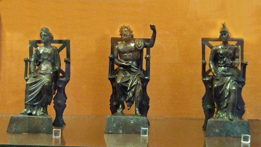 Three small bronze statues seated on thrones, found on the top step of the lararium of VI.16.7.  
Juno (left), Jupiter in the centre and Minerva (right).  
On the same step, to the left of Juno, stood the statue of Mercury. 
The two Lares were placed, on either side of the group, but on the lower step.
Now in Naples Archaeological Museum. Jupiter is inventory number 133323, Juno is 133324 and Minerva is 133325.
See Boyce G. K., 1937. Corpus of the Lararia of Pompeii. Rome: MAAR 14, pp. 57-8, no. 221, and pl.38, 2
