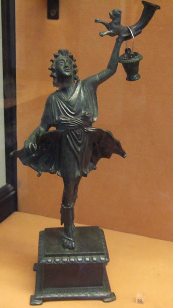 Bronze statuette of a Lar, found in Lararium of VI.16.7. 
This was one of two Lares that were found on the lower step of the lararium.
The Lar holds a rhyton which has at its lower end the front half of a panther.
The bucket would normally be in the Lar’s right hand.
Now in Naples Archaeological Museum. Inventory number 133327.
See Notizie degli Scavi di Antichità, 1907, p. 567, fig. 17.

