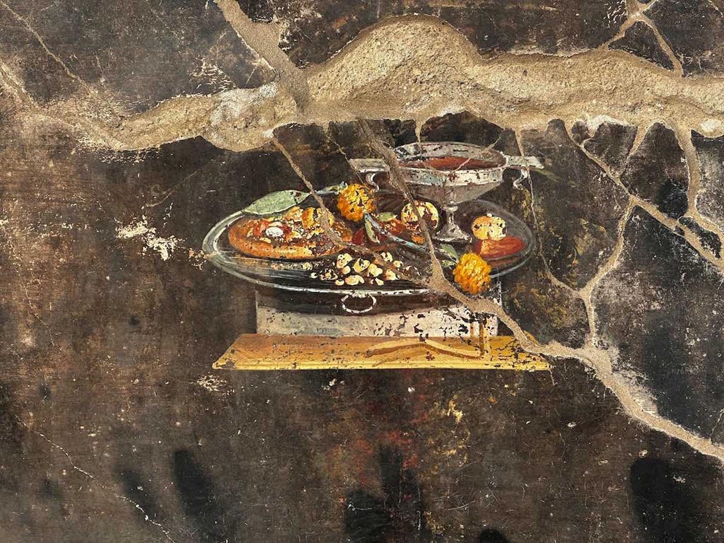 IX.10.1 Pompeii. June 2023. Atrium 2 west wall. It looks like a pizza?
Whilst it looks like a pizza, this image from a Pompeian painting from 2000 years ago obviously can't be, since some of the most characteristic ingredients are missing, namely tomatoes and mozzarella. However, it could be a distant ancestor of the modern dish, elevated to the status of a World Heritage element in 2017 as the "traditional art of the Neapolitan pizza chef'.
As the archaeologists of the Archaeological Park of Pompeii explain, it is possible to suppose that next to a wine cup placed on a silver tray, there is depicted a flat focaccia that functions as a support for various fruits, (that can be identified as pomegranate, and maybe a date), with spices and perhaps with a type of pesto, (moretum in Latin) as indicated by yellow and ochre dots, possibly condiments. Moreover, on the same tray, dried fruits and a garland of yellow strawberry trees are present next to the dates and pomegranate.
See PAP: Pompeii: a still life discovered by the new excavations of Regio IX

IX.10.1 Pompeii. Giugno 2023. Atrio 2 parete ovest. Sembra una pizza?
Sembra una pizza, quello che si vede su un dipinto pompeiano di 2000 anni fa, ma ovviamente non lo può essere, a rigore, dato che mancavano alcuni degli ingredienti più caratteristici, ovvero pomodori e mozzarella. Tuttavia potrebbe essere un lontano antenato della pietanza moderna, elevata a patrimonio dell'umanità nel 2017 in quanto "arte tradizionale del pizzaiuolo napoletano'.
Come spiegano gli archeologi del Parco Archeologico di Pompei, si suppone che accanto a un calice di vino, posato su un vassoio di argento, sia raffigurata una focaccia di forma piatta che funge da supporto per frutti vari (individuabili un melograno e forse un dattero), condita con spezie o forse piuttosto con un tipo di pesto (moretum in latino), indicato da puntini color giallastro e ocra. Inoltre, presenti sullo stesso vassoio, frutta secca e una ghirlanda di corbezzoli gialli, accanto a datteri e melograni.
Photograph courtesy of Davide Peluso.
Vedi PAP: Pompei: emerge una natura morta dai nuovi scavi della Regio IX
