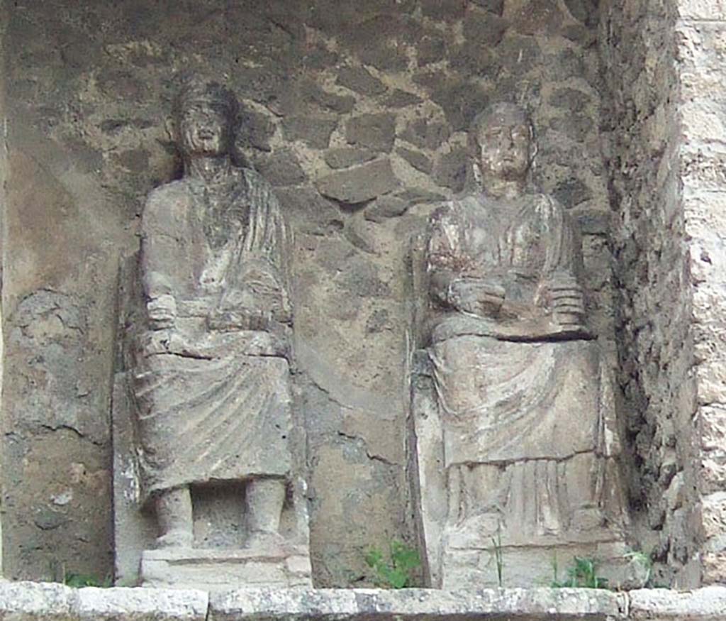 Pompeii Porta Nocera. May 2006. Tomb 9OS. Statues of the seated couple in the tomb.
The couple are destined to remain anonymous as there are no inscriptions.
The roll in the hand of the male character would suggest a function of magistrate who would be well in keeping with the dignity of the monument.
See D’Ambrosio, A. and De Caro, S., 1983. Un Impegno per Pompei: Fotopiano e documentazione della Necropoli di Porta Nocera. Milano: Touring Club Italiano. (9OS)


