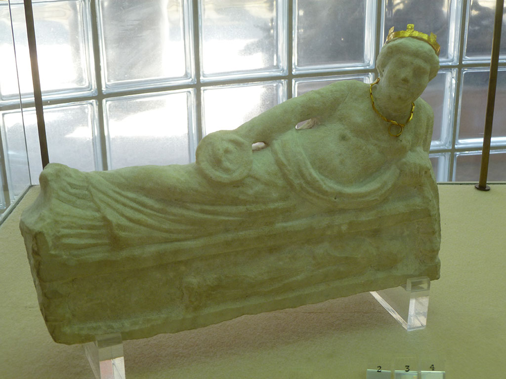 Boscoreale, Villa rustica in via Casone Grotta, proprietà Risi di Prisco. Lararium. 
Marble statuette of a female Genius, relaxing on a couch (kline), dressed in chiton and cloak.
The gold ring, necklace and diadem that decorate it were found beside it.
Now in Boscoreale Antiquarium. Photo courtesy of Michael Binns.
See Fergola, L., 1987. Comune di Boscoreale. Via Casone Grotta, proprietà Risi Di Prisco, in Rivista di Studi Pompeiani, 1, p. 164.
The objects on display in the Boscoreale Antiquarium were found when the villa’s private sacellum (shrine) was excavated. 
A small statue of a female deity at a banquet, identifiable as Bona Dea. The simulacrum was decorated with golden miniature jewellery: a tiara and a necklace with a small ring hung on it.
http://pompeiisites.org/en/uncategorized/antiquarium-room-ii/ 
