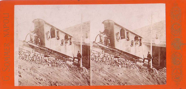 Vesuvius Funicular railway at the lower station, 1880-1889. One of the two original 1880 cars named Etna and Vesuvio which seated 8 people.
There was a longitudinal wooden sleeper on the top of which was carried a single rail. 
The cars had a double flanged wheel at each end which ran on this rail. 
In addition, there were two angled rails, one fixed to each side of the sleeper at its base. 
The cars had wheels mounted from their floors which engaged on these side rails and kept the cars upright. 
Each track had two continuous cables carried on pulleys which were fixed to each side of the car.
These cars were replaced in 1889 by new cars with 10 seats, also named Etna and Vesuvio, as part of renovations by John Mason Cook.
Stereoview photo by Giorgio Sommer.
