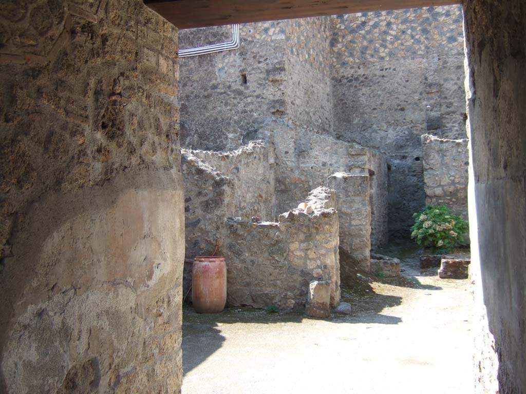 I.10.18 Pompeii. September 2005. Looking south-west from entrance fauces across atrium.
Right of centre is the area of room 6, looking ahead to cistern mouth in walkway No.8.
On the left at the end of the entrance corridor wall is the south-west side of the atrium.
Left of centre is room 7, the kitchen (room 9) is at the rear of room 7.

According to Boyce, in the south wall of the kitchen beside the hearth was a fragmentary painting in two zones.
It was on a white background outlined with red stripes.
In the upper zone stood the Genius, with a Lar.
In the lower zone, beneath garlands, was the tail of a black and yellow serpent.
Below the serpent were several kitchen articles, a hog’s head, a ham on a nail, and an eel on a spit.
See Notizie degli Scavi di Antichità, 1934, p. 344, and fig.38.
See Boyce G. K., 1937. Corpus of the Lararia of Pompeii. Rome: MAAR 14. (p.29, no.60) 
Fröhlich described it was “mostly fallen from the wall, and only the body of the left Lar is quite well preserved.”
See Fröhlich, T., 1991. Lararien und Fassadenbilder in den Vesuvstädten. Mainz: von Zabern. (L18, Taf. 26, 3).
