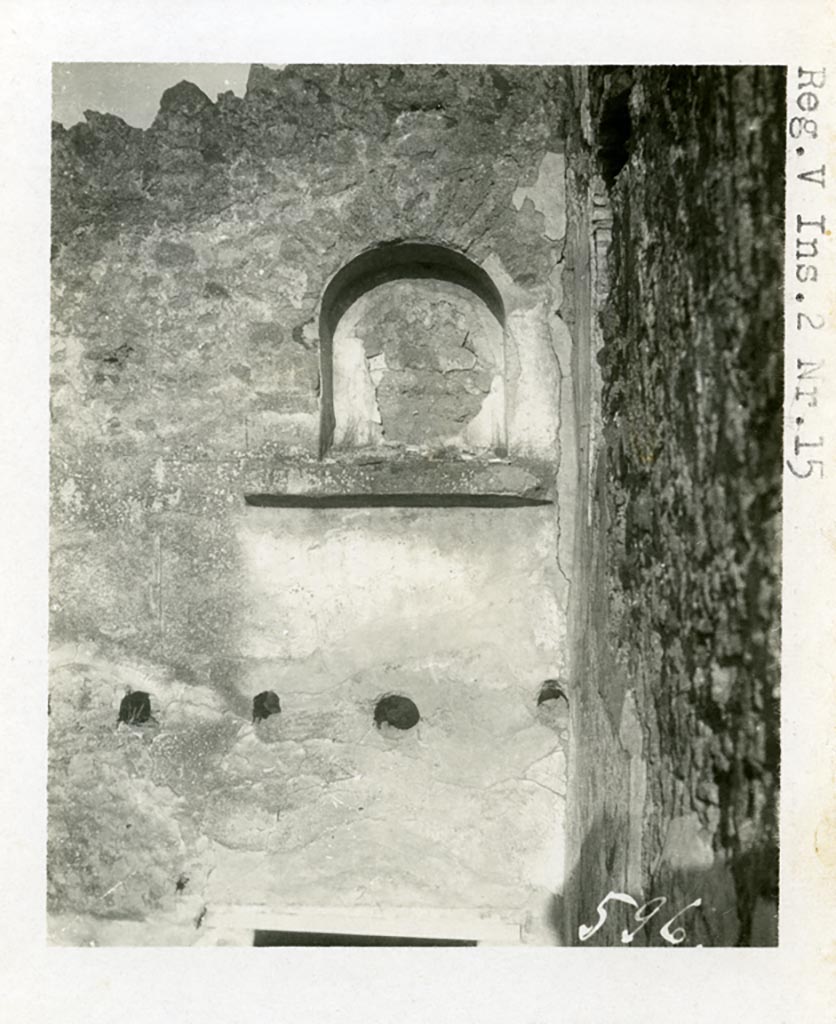 V.2.d Pompeii but shown as V.2.15 on the photo. Pre-1937-39. Niche “n” on plan.
Looking towards vaulted niche lararium on upper north wall, in north-east corner of rear of house. 
Photo courtesy of American Academy in Rome, Photographic Archive. Warsher collection no. 596
According to Boyce, there were two lararia in the house.
One on the north wall of a rear room that occupied the place of the peristyle, high above the floor, was a large arched niche (0.8m high, 0.65m wide, 0.35m deep, 2.9m high above the floor) with a heavy projecting ledge beneath it. 
Painted on the back wall of this niche, was a burning altar adorned with a garland and furnished with two eggs, and on each side of the altar was a palm branch, yellow with red ribbon attached. 
The wall around the niche was covered with white stucco which below the niche was studded with red spots representing a dado, down as far as a row of holes, where a kind of mezzanine was attached, by means of which the niche was reached. 
At a late date this structure was removed and Mau supposed that the niche was reached thereafter by a portable ladder.
See Boyce G. K., 1937. Corpus of the Lararia of Pompeii. Rome: MAAR 14. (p.36, nos.101 and 102,)
See Bullettino dell’Instituto di Corrispondenza Archeologica (DAIR), 1885, p.256.
According to PPM, nothing remains of the painting other than a small amount of the base stucco.
