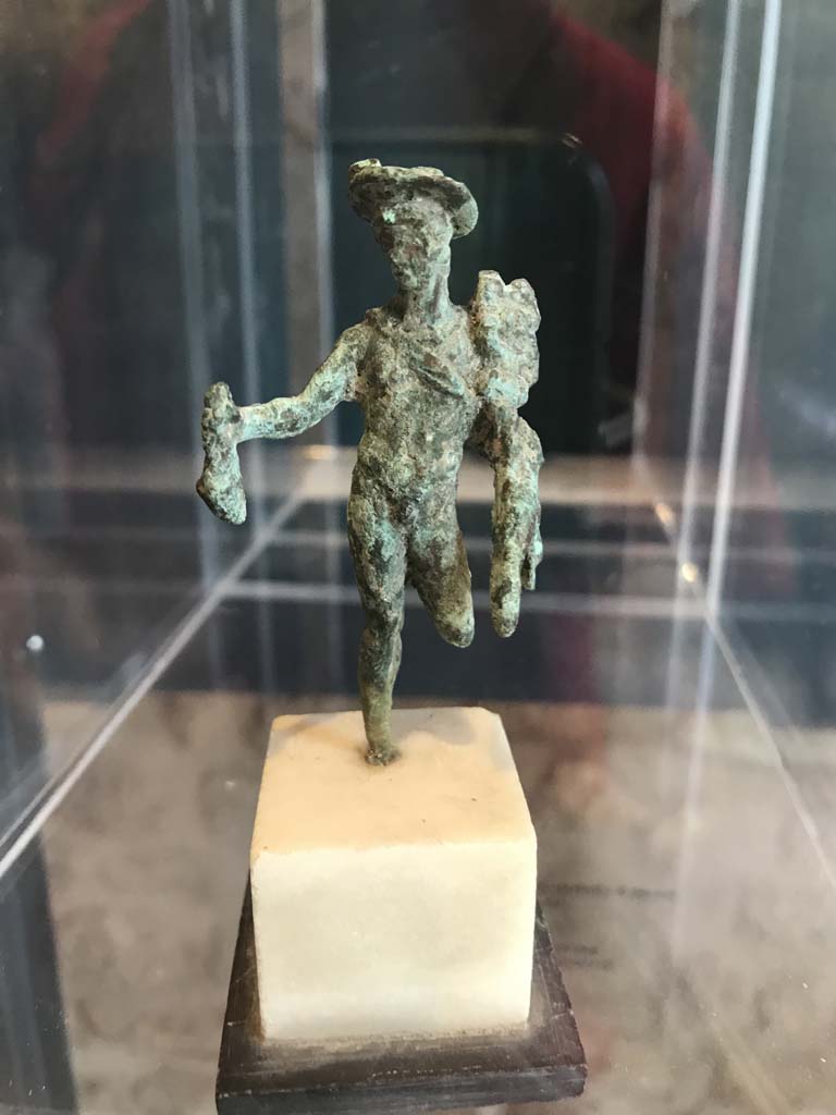 VIII.2.1 Pompeii. April 2019. Bronze statuette of Mercury carrying a money bag.
Found in the area to the south of the House of Championnet.
Pompeii Inventory number 6331. Photo courtesy of Rick Bauer.
According to Zanella -
This statuette was found in Room B, the triclinium on the lower floor of VIII.2.3-5 (or VIII.2.14-16) on 9th February 1937.

9/2/1937
In room B an imprint of a box had been removed at a distance of m. ,0,70 from the bed frame, this imprint had been found to be m.0.68 x 0.50 x 0.22 including a shelf in the front m.0.50 x 0.18 x 0.16.
In removing the last clod of ash with the partial imprint of the triclinium couch, the following was found: Bronze: statue representing Mercury standing missing a (leg) with the pouch and caduceus; it was fragmented and missing its left leg from the knee down and the right foot. 
Alt m.0.068 (Pompeii inv. 6331)
(Nel ambiente B si era rimosso un’impronta di cassa distante m. ,0,70 dal telaio del letto, detta impronta era risultata di m.0.68 x 0.50 x 0.22 compreso un ripiano nella parte anteriore m.0.50 x 0.18 x 0.16.
Nel rimuovere l’ultima zolla di cenere con l’impronta parziale del letto tricliniare si era rinvenuto: bronzo: statuina rappresentante Mercurio stante mancante di un (*) col marsupio e caducco; era frammentata e mancante della gamba sinistra dal ginocchio in giù e del piede destro. Alt m.0.068 (Pompei inv. 6331))
See Zanella S., 2019. La caccia fu buona: Pour une histoire des fouilles à Pompéi de Titus à l’Europe. Naples : Centre Jean Bérard, p. 268.
