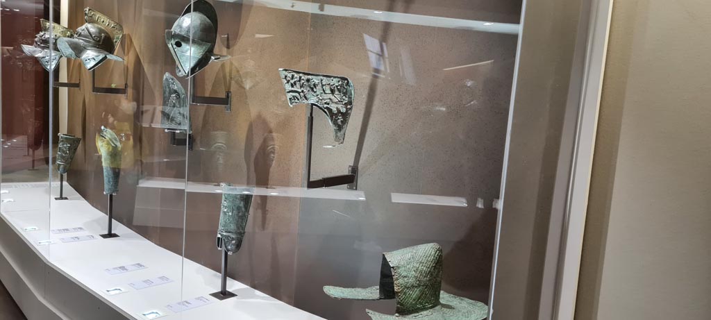 Items in display cabinet in Naples Archaeological Museum. Photo taken May 2021, courtesy of Giuseppe Ciaramella.
On far left –Helmet and greave, inv. 5671 and 5676 (from Pompeii VII.11.6). 
Then – helmet with relief of head of Medusa
Then – Secutor’s helmet, inv. 5642. 
Then – (from Herculaneum) crest of Murmillo’s helmet with depictions of Mars, Rhea Silvia, she-wolf with Romulus and Remus and deities, found 19th June 1834. Naples Archaeological Museum, inv. 5656.
Lower right - Bronze Pileus (pilos helmet) inv. 5651.
