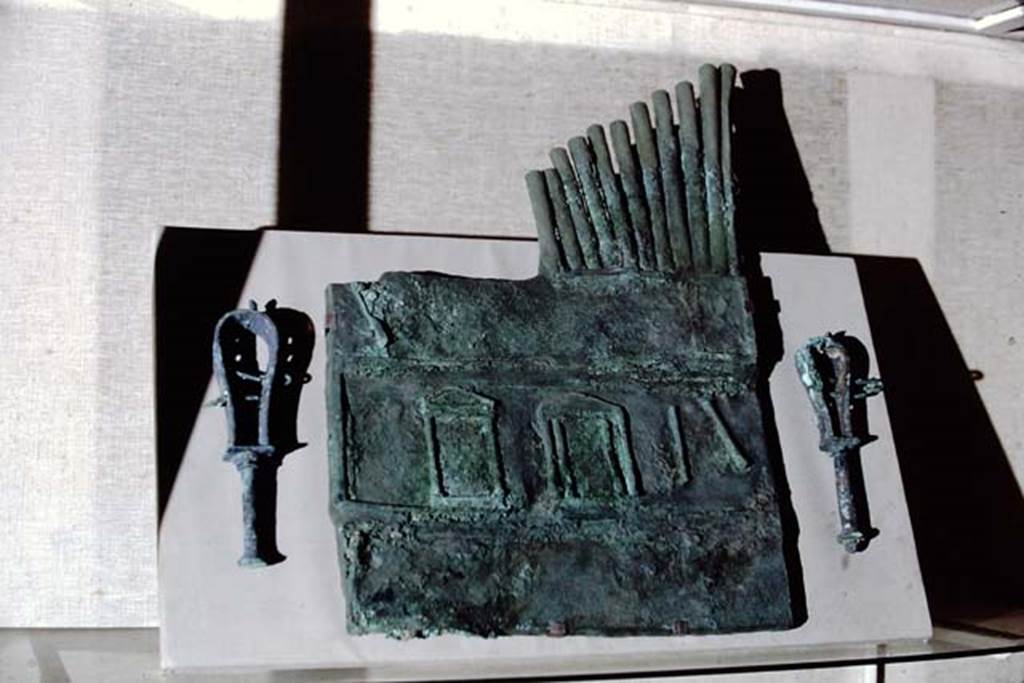 Villa of T. Siminius Stephanus, fondo Barbatelli. 1968. Bronze Pan pipes (syrinx)discovered in 1899.
The Pan pipes are decorated with three aediculae that are supposed to represent a frons scaenae and, at the top, nine rods of different heights.
They are connected at the bottom, and bear a hole near of the mouth. 
The object, strongly restored in the nineteenth century, is large, so as to suggest that the operation needed the help of a special machine. 
Its use was connected to the satyr play and the myth, but the instruments were also used during ritual ceremonies, such as Isiac processions, during which the faithful waved the sistrum.
It was also used during theatrical performances to entertain banqueters, as music as a separate discipline from acting does not seem to have existed in ancient times.
Now in Naples Archaeological Museum. Inventory number 125187 but shown on the MANN web site as 111055.
Photo by Stanley A. Jashemski.
Source: The Wilhelmina and Stanley A. Jashemski archive in the University of Maryland Library, Special Collections (See collection page) and made available under the Creative Commons Attribution-Non-Commercial License v.4. See Licence and use details.
J68f1410

