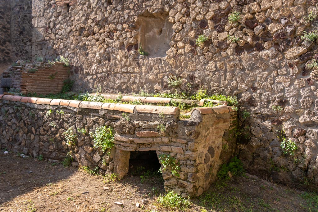 Villa of Mysteries, Pompeii. October 2023. 
Room 61, west wall with hearth and oven, and remains of niche. Photo courtesy of Johannes Eber.
According to NdS, another niche lararium was above the podium of the hearth and oven.
The stucco had fallen and brought to light a part of an ancient painted lararium, for all its simplicity, not without interest.
At the side were the remains of a painted helmeted Minerva, and of another divinity, perhaps Vulcan.
See Notizie di Scavi, 1910, p.141-2.
According to Boyce –
In the east wall of the same courtyard, above the hearth, is a second rectangular niche (h.070, w.0.60, d.0.20) on the back wall of which De Petra saw painted the figures of Minerva armed with the helmet and of another divinity – perhaps Vulcan.
Reference Maiuri, Villa dei Misteri, 81.
See Boyce G. K., 1937. Corpus of the Lararia of Pompeii. Rome: MAAR 14. (p.98, no. 482).

